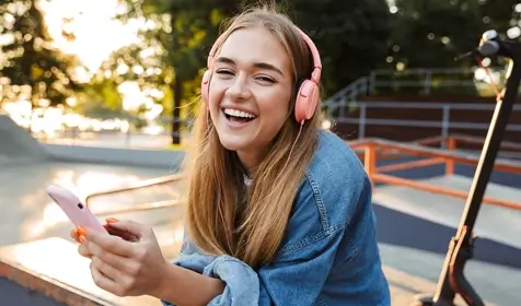 Image of a laughing cheery smiling young teenage girl outside in park listening music with headphones holding mobile phone showing tongue.